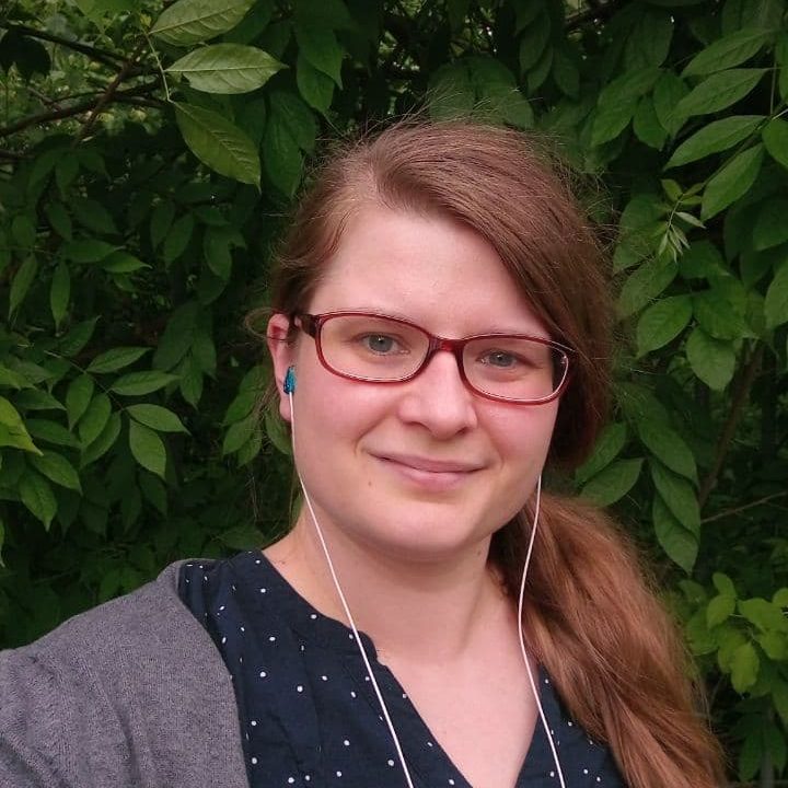 Farbfoto einer weißen Frau mit dunkelblonden Haaren und Brille vor grünen Bäumen